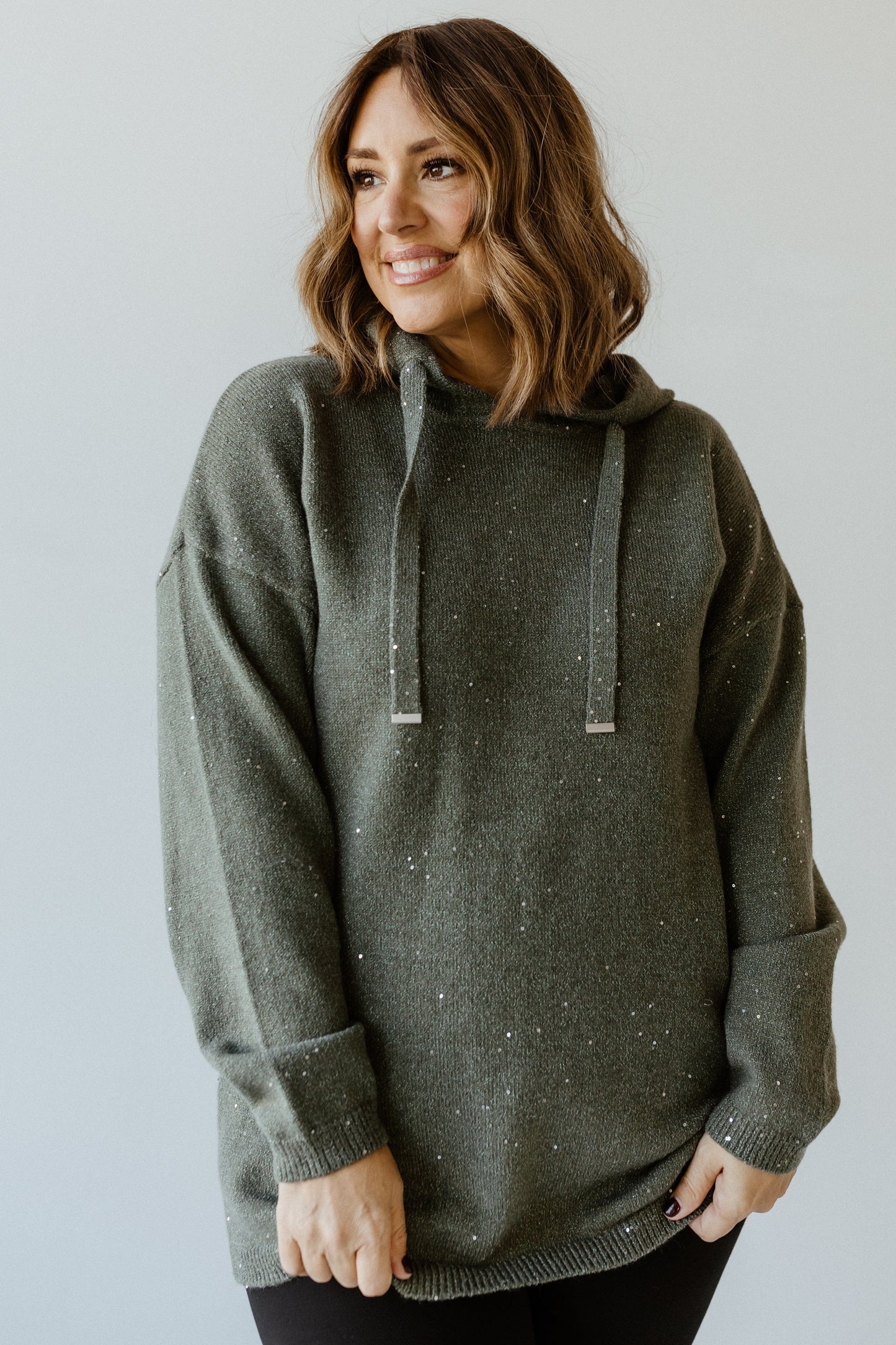 A person with shoulder-length hair is wearing a dark green speckled hoodie with silver toggles and black pants, standing against a light background, smiling and looking to the side.