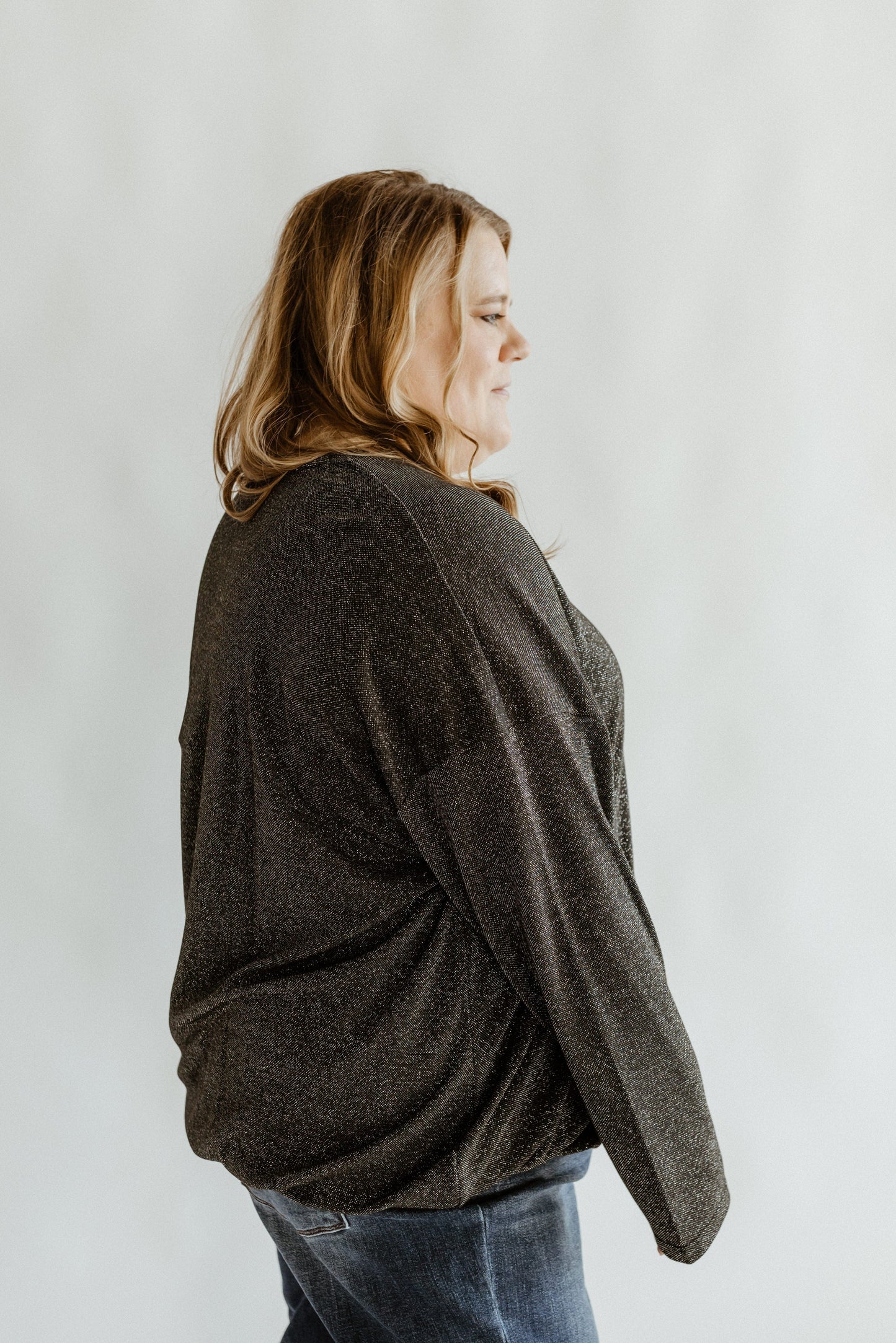 A person with long hair stands sideways, showcasing the luxurious fabric of a dark sweater and jeans, against a plain light background.