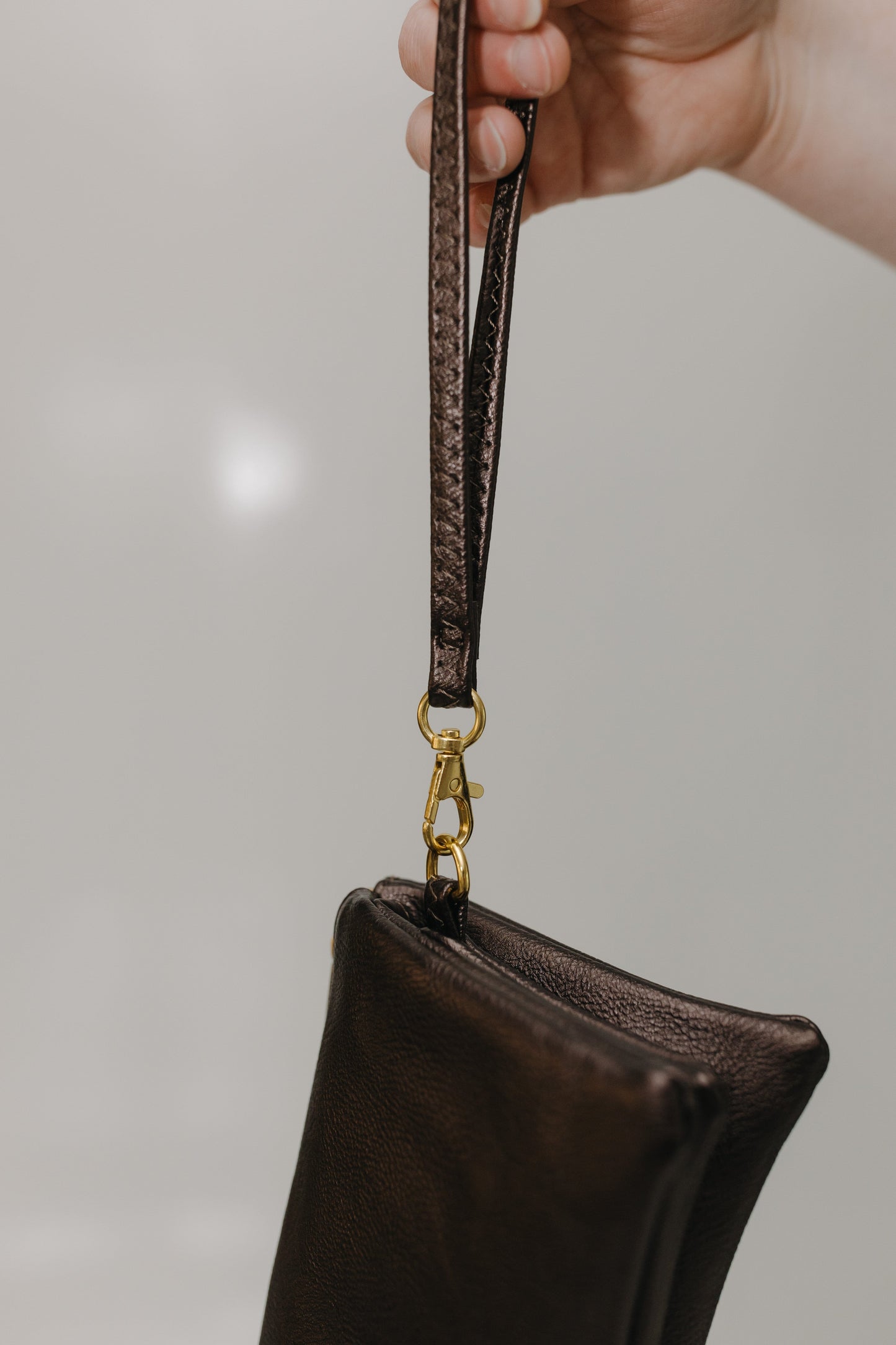 Brown leather pouch with a strap held by a hand against a neutral background