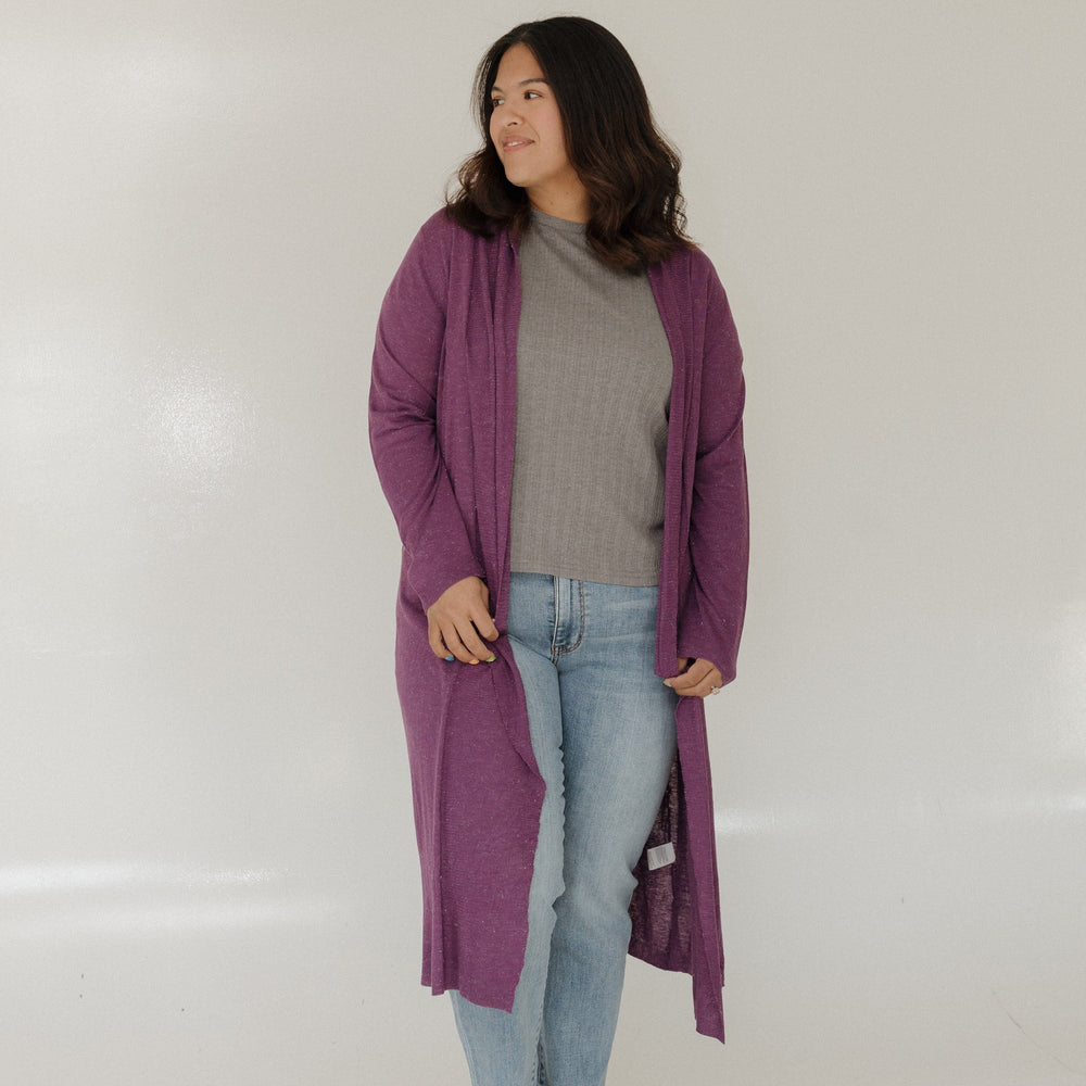 Woman wearing a long purple cardigan, gray sweater, blue jeans, and beige shoes on a white background