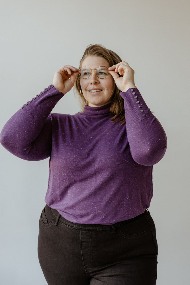 A person wearing Yesta's SHIMMERY LIGHTWEIGHT TURTLENECK IN ROYALTY and glasses stands against a plain background, adjusting their glasses with both hands.