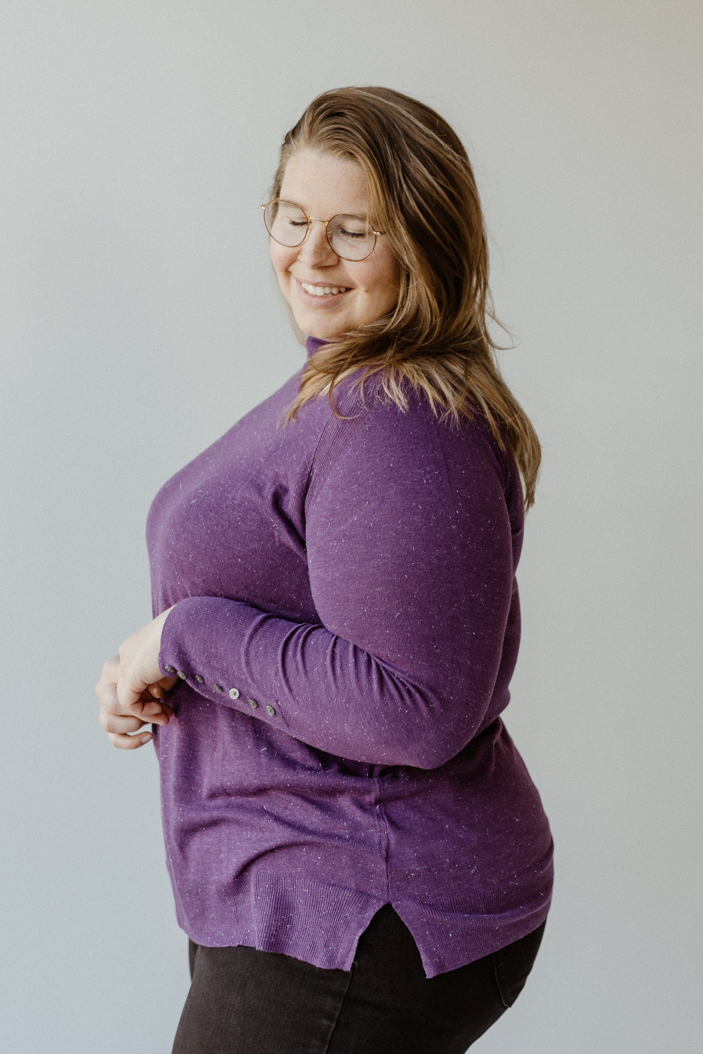 A woman with long hair and glasses, wearing Yesta's shimmery lightweight turtleneck in royalty paired with dark pants, stands against a plain background, smiling and looking down.