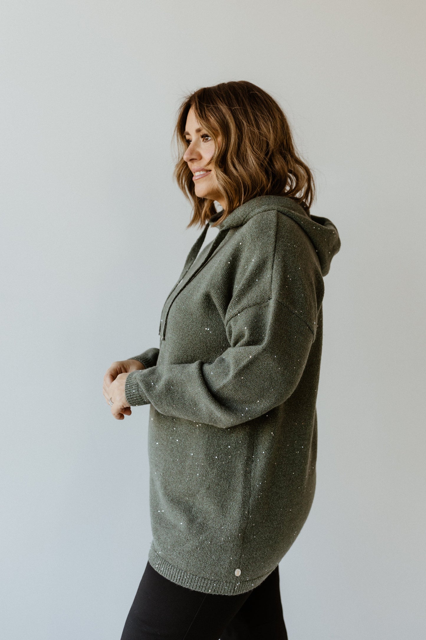 A woman in a green sparkly hoodie with silver toggles and black pants stands sideways against a light background.