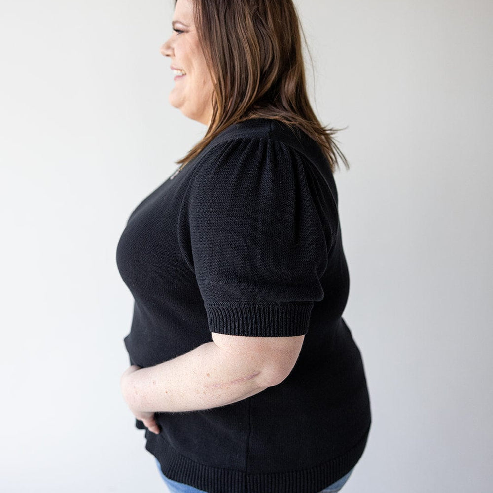 A woman with brown hair is standing in profile, wearing a stylish SHORT PUFF SLEEVE SWEATER from Love Marlow and blue jeans, smiling against a plain white background. Her preppy knit choice adds a touch of elegance to her versatile wardrobe.