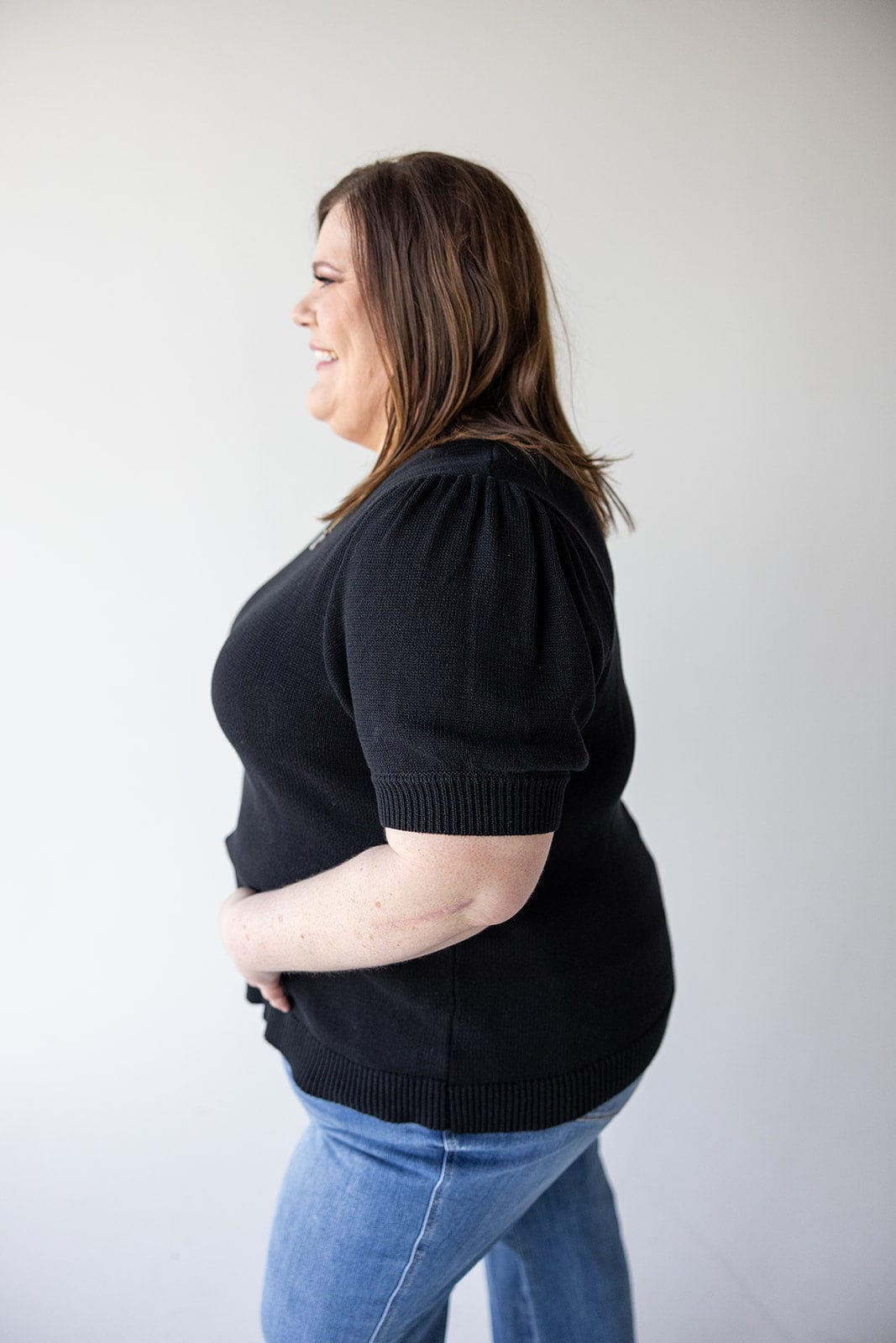 A woman with brown hair is standing in profile, wearing a stylish SHORT PUFF SLEEVE SWEATER from Love Marlow and blue jeans, smiling against a plain white background. Her preppy knit choice adds a touch of elegance to her versatile wardrobe.