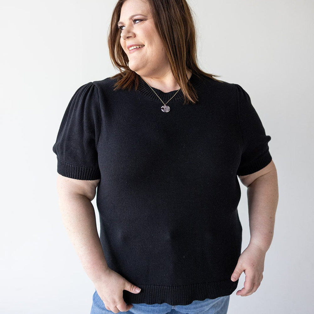 A person with shoulder-length brown hair stands against a plain background, wearing a Love Marlow SHORT PUFF SLEEVE SWEATER and blue jeans, smiling and looking to the side. Their versatile wardrobe choice adds an effortless charm to their look.