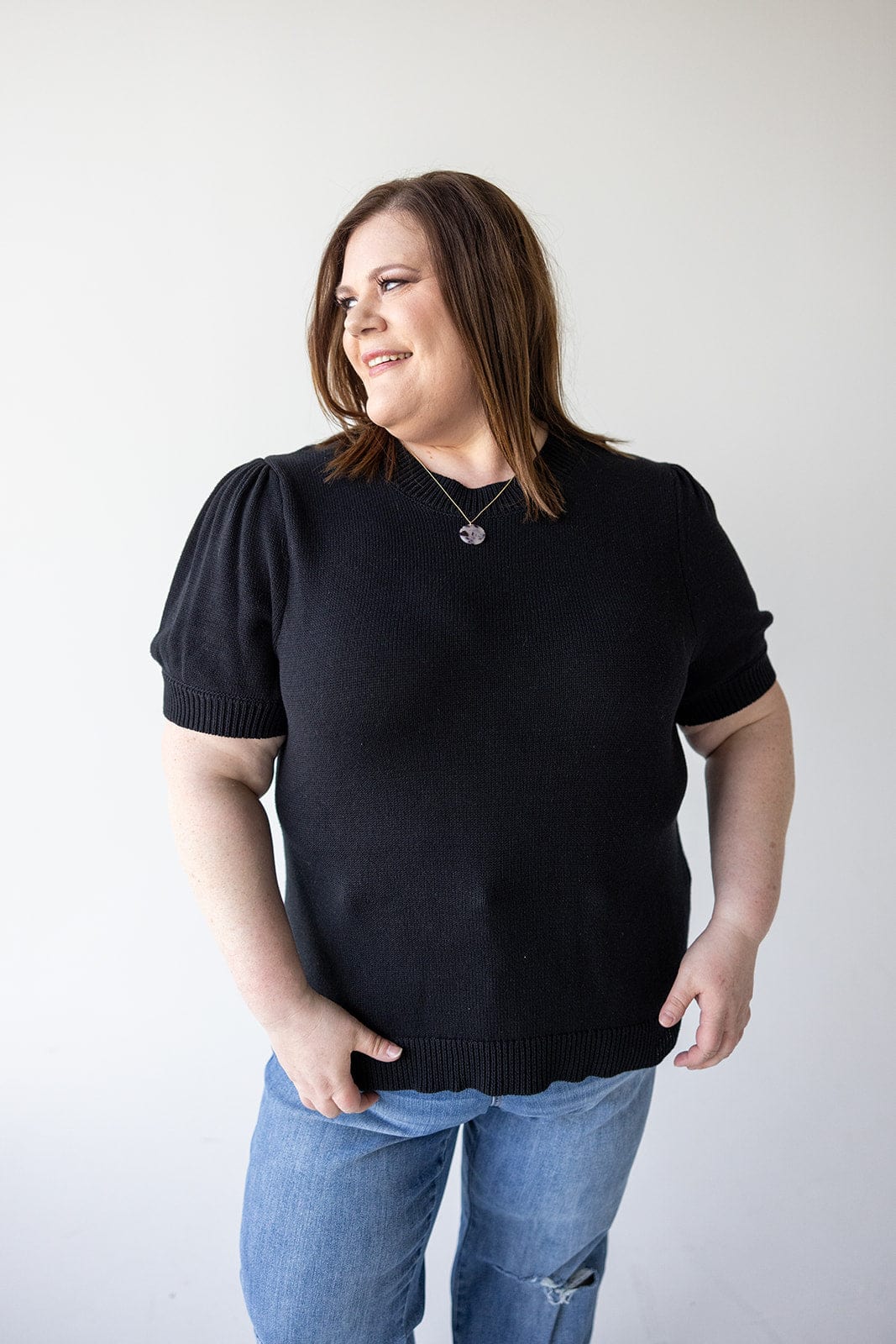 A person with shoulder-length brown hair stands against a plain background, wearing a Love Marlow SHORT PUFF SLEEVE SWEATER and blue jeans, smiling and looking to the side. Their versatile wardrobe choice adds an effortless charm to their look.