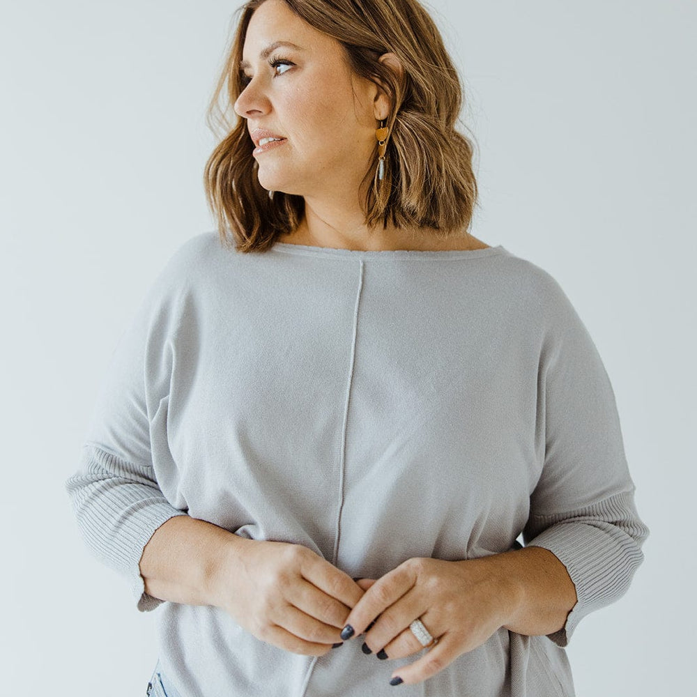 A woman with shoulder-length hair is standing in a SHORT SLEEVE SEAM SWEATER IN DOVE by Love Marlow and jeans, looking to the left, with her hands lightly clasped in front of her, exuding a sophisticated style.