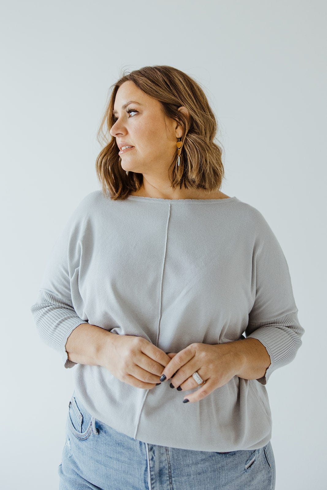 A woman with shoulder-length hair is standing in a SHORT SLEEVE SEAM SWEATER IN DOVE by Love Marlow and jeans, looking to the left, with her hands lightly clasped in front of her, exuding a sophisticated style.