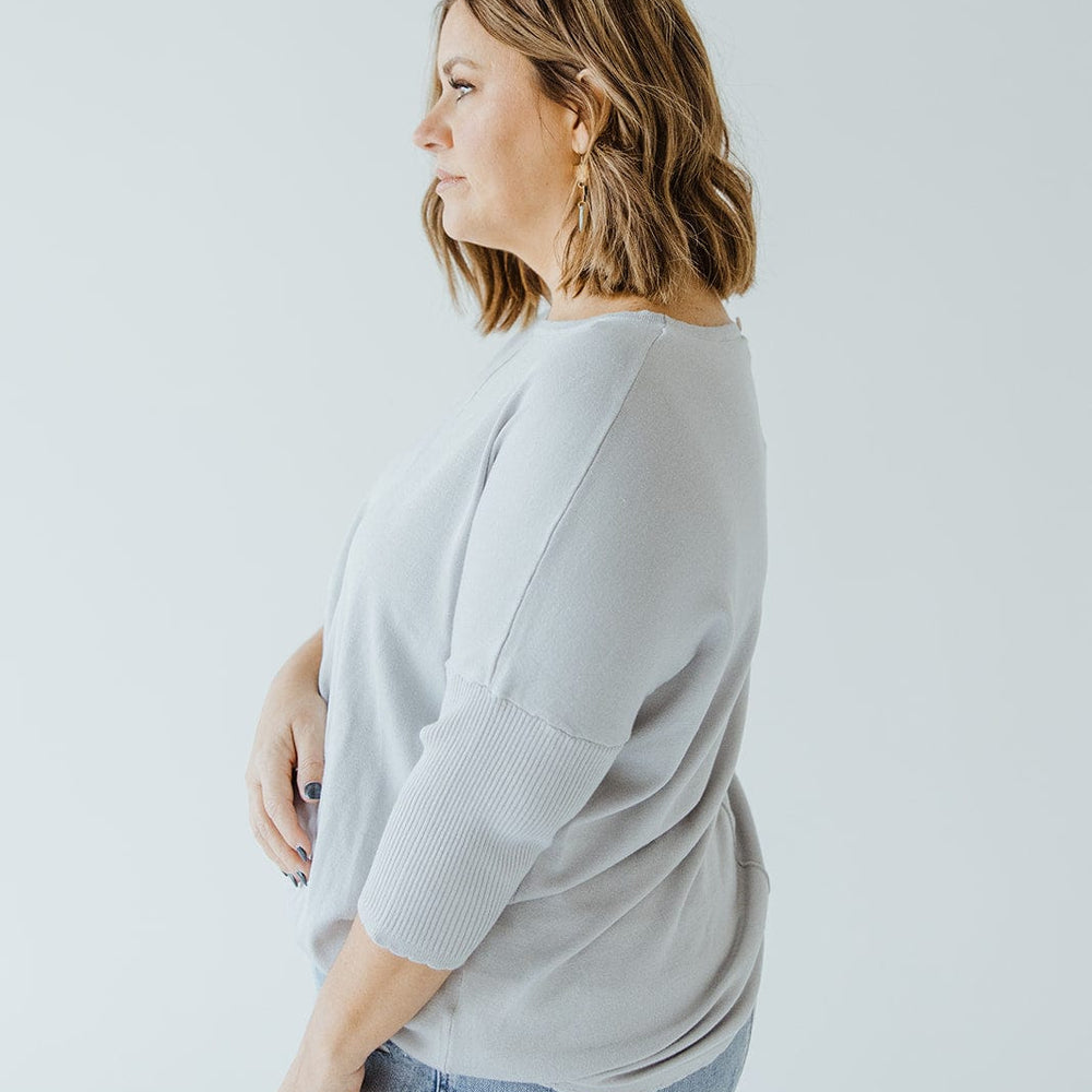 A woman with shoulder-length hair is standing in profile view, wearing a SHORT SLEEVE SEAM SWEATER IN DOVE by Love Marlow and light blue jeans against a plain white background, embodying a sophisticated style.