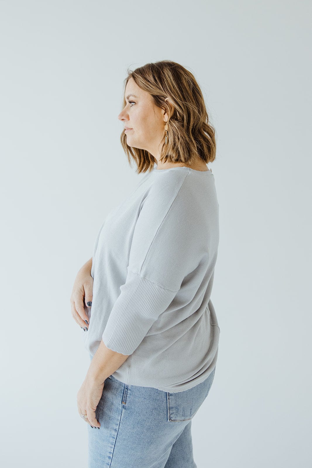 A woman with shoulder-length hair is standing in profile view, wearing a SHORT SLEEVE SEAM SWEATER IN DOVE by Love Marlow and light blue jeans against a plain white background, embodying a sophisticated style.