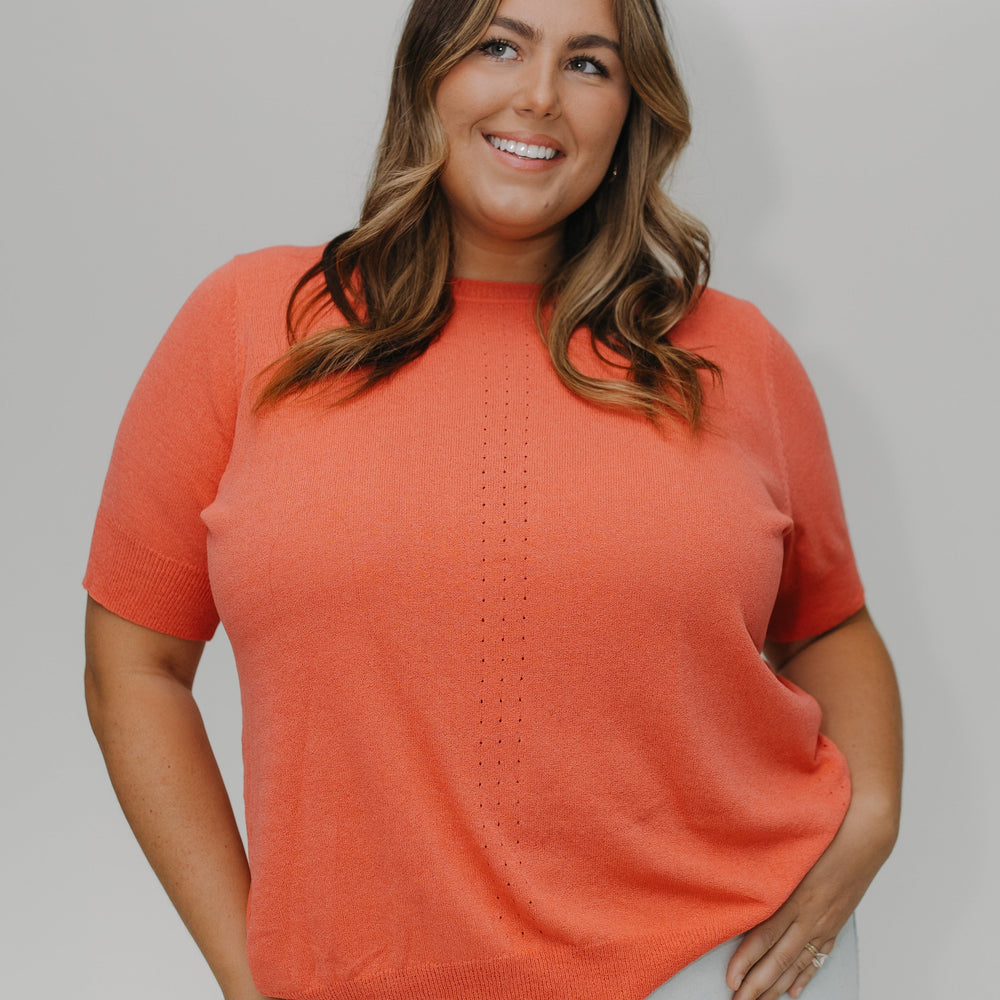 Woman wearing a coral sweater and light jeans against a plain background