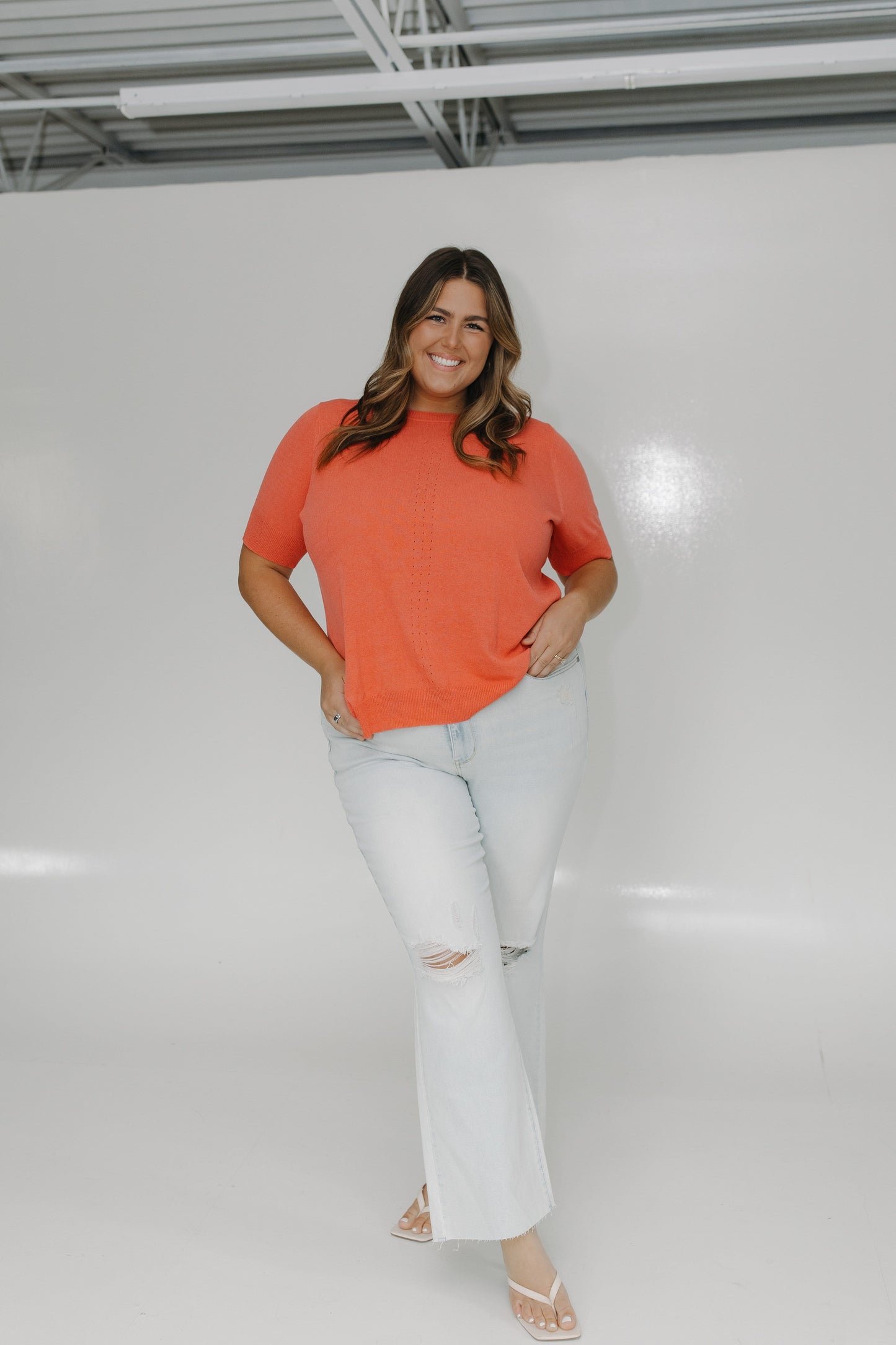 Woman wearing an orange shirt and white jeans standing against a plain white background