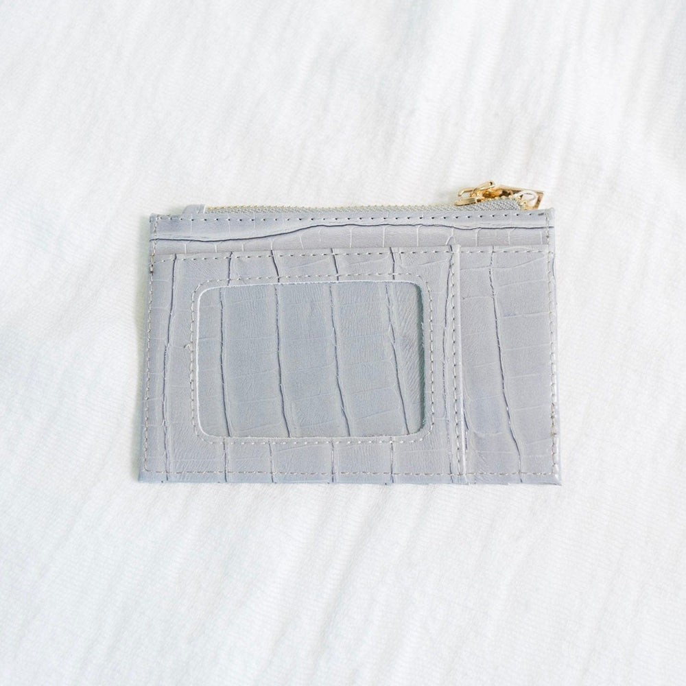 A smoky croc wallet crafted from faux leather rests on intertwined wooden branches, with a green potted plant in the background. This sleek cardholder is RFID protected, offering both style and security in one elegant package.