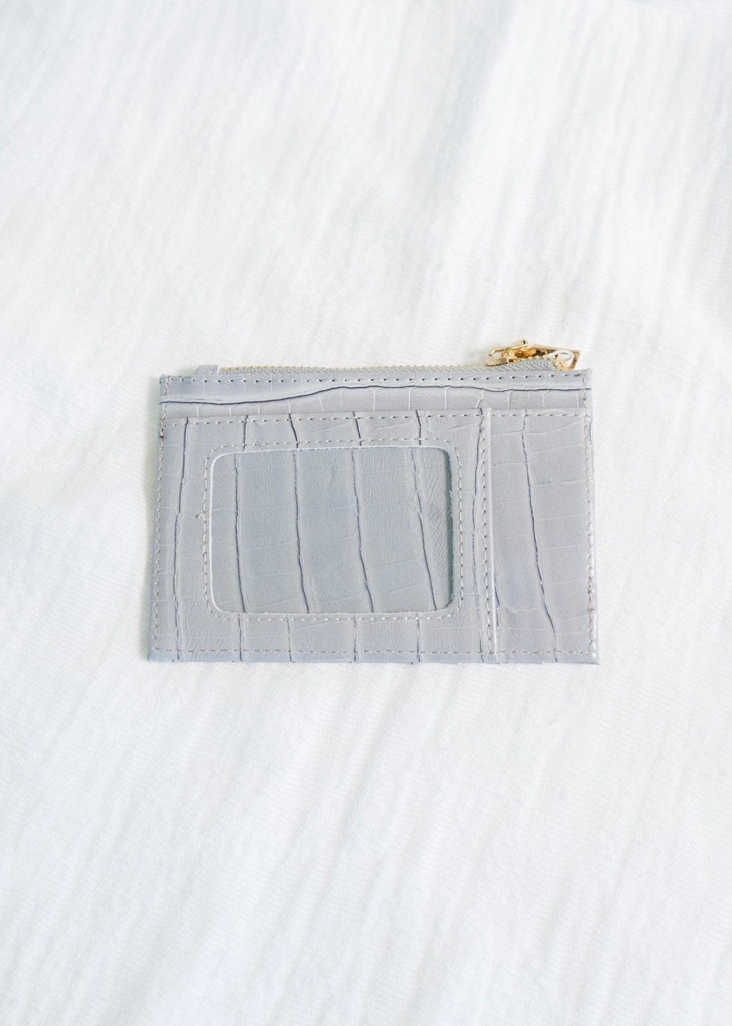 A smoky croc wallet crafted from faux leather rests on intertwined wooden branches, with a green potted plant in the background. This sleek cardholder is RFID protected, offering both style and security in one elegant package.