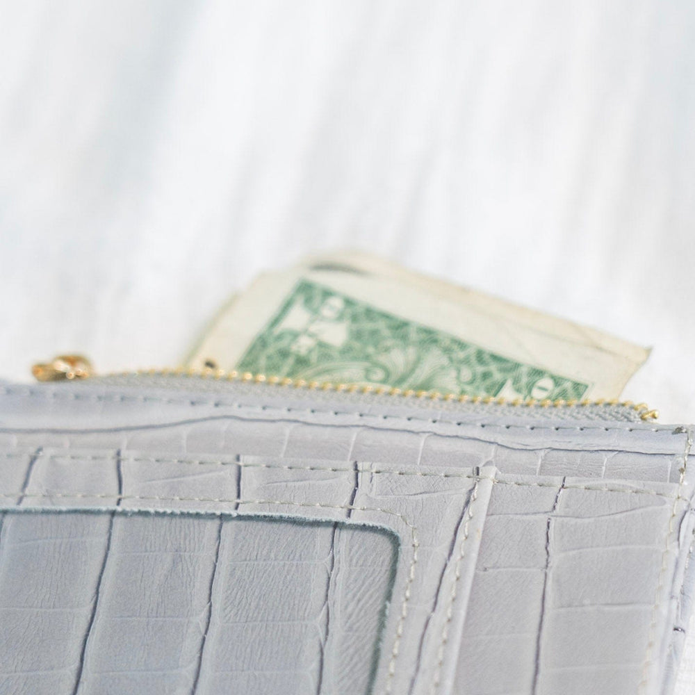 A smoky croc wallet crafted from faux leather rests on intertwined wooden branches, with a green potted plant in the background. This sleek cardholder is RFID protected, offering both style and security in one elegant package.