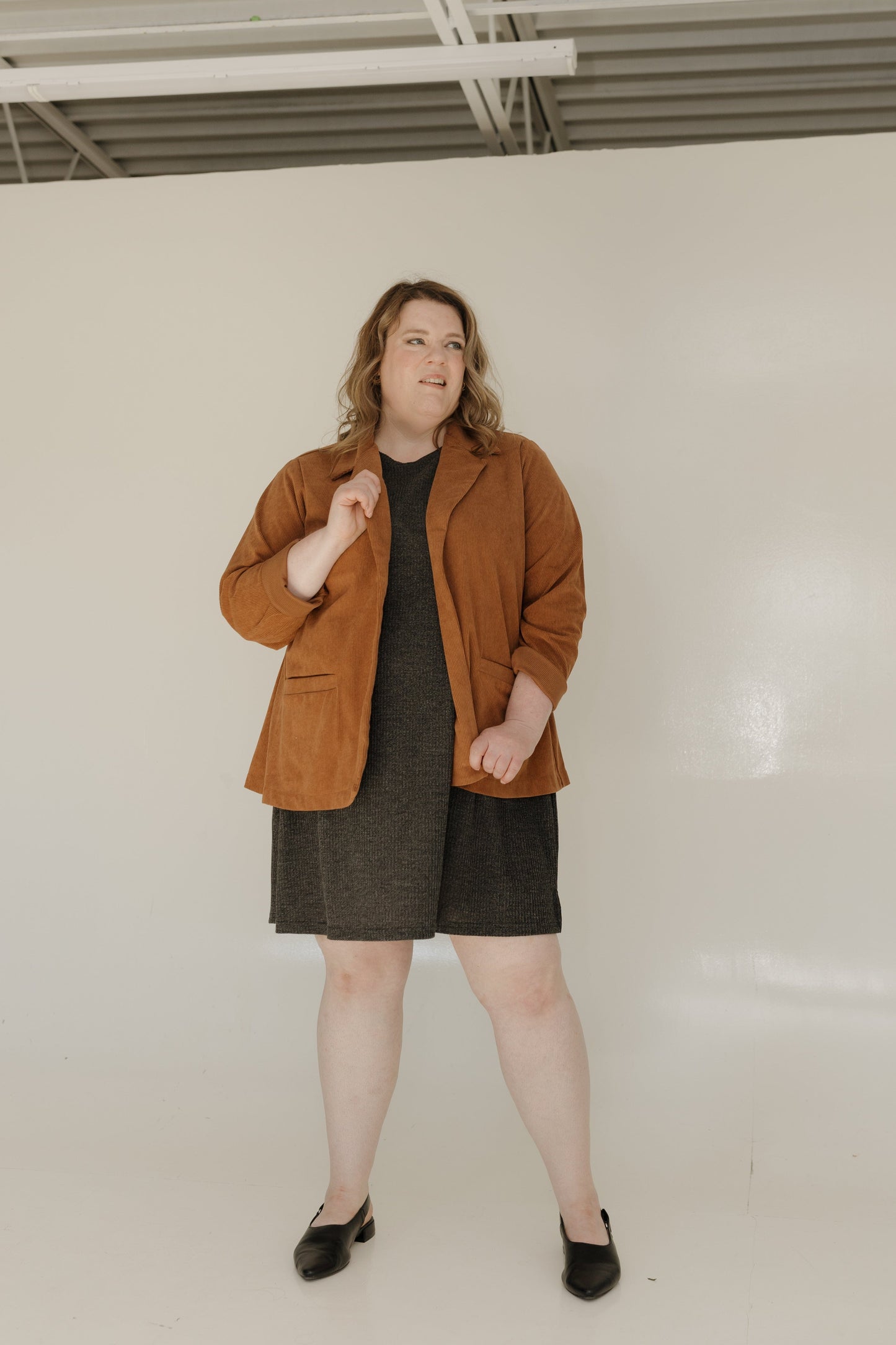 Woman wearing a brown jacket over a black dress against a white wall