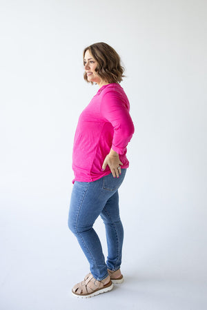 A woman in a bright pink 3/4 Sleeve Tunic with Button Detail in Glam Pink by Love Marlow and blue jeans stands in a side profile pose against a white background.