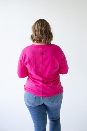 A person with shoulder-length hair wearing a bright pink 3/4 SLEEVE TUNIC WITH BUTTON DETAIL IN GLAM PINK from Love Marlow and blue jeans stands facing away from the camera against a plain white background.