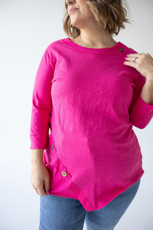 A woman wearing a bright pink, 3/4 SLEEVE TUNIC WITH BUTTON DETAIL IN GLAM PINK by Love Marlow, paired with blue jeans, poses with one hand near her shoulder and the other relaxed by her side.