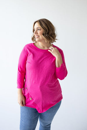 A person with shoulder-length hair, wearing a bright pink **3/4 SLEEVE TUNIC WITH BUTTON DETAIL IN GLAM PINK** from **Love Marlow** and blue jeans, stands against a plain white background.