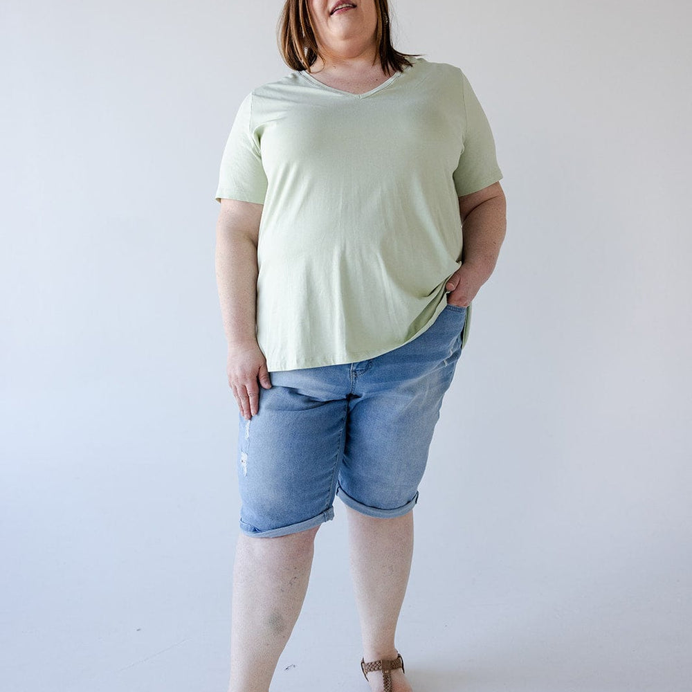 A woman with medium-length brown hair wearing a light green t-shirt, Love Marlow SLIGHTLY DISTRESSED CUFFED BERMUDA SHORT, and sandals stands against a plain white background. She has one hand in her pocket.