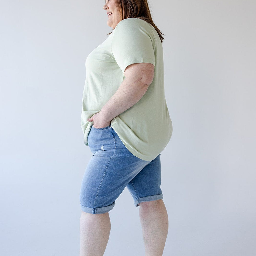 Person standing sideways wearing a light green t-shirt, Love Marlow SLIGHTLY DISTRESSED CUFFED BERMUDA SHORT with five-pocket construction for a stylish yet comfortable fit, and sandals, against a plain white background.