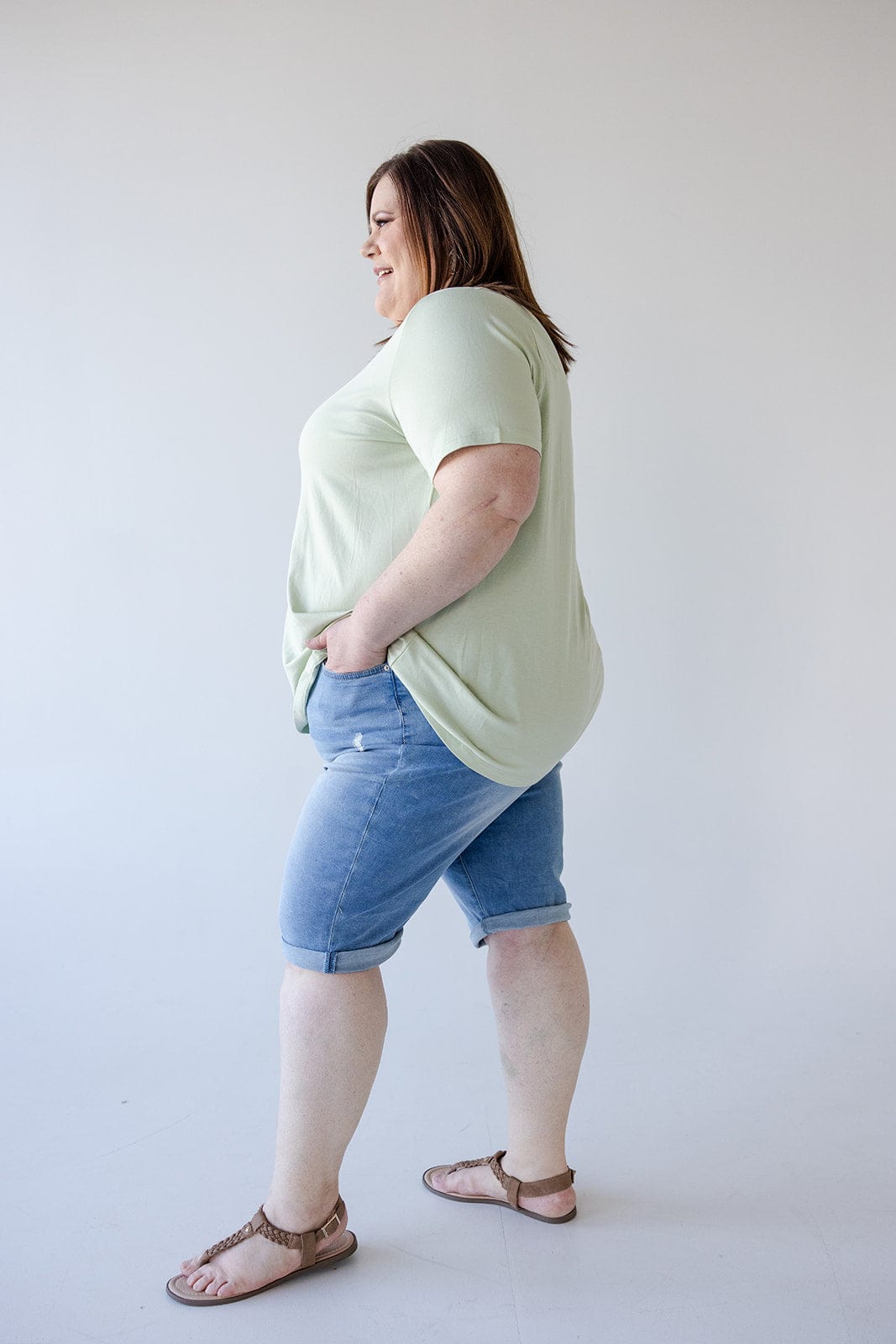 Person standing sideways wearing a light green t-shirt, Love Marlow SLIGHTLY DISTRESSED CUFFED BERMUDA SHORT with five-pocket construction for a stylish yet comfortable fit, and sandals, against a plain white background.