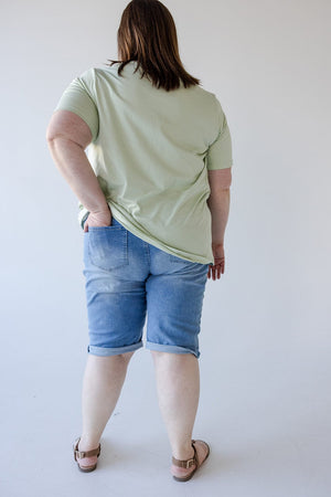 A person with light skin, seen from behind, wearing a green T-shirt, Love Marlow SLIGHTLY DISTRESSED CUFFED BERMUDA SHORT for a comfortable fit, and sandals, standing against a plain white background.