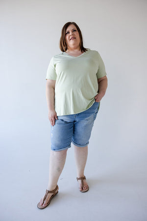 A woman with medium-length brown hair wearing a light green t-shirt, Love Marlow SLIGHTLY DISTRESSED CUFFED BERMUDA SHORT, and sandals stands against a plain white background. She has one hand in her pocket.