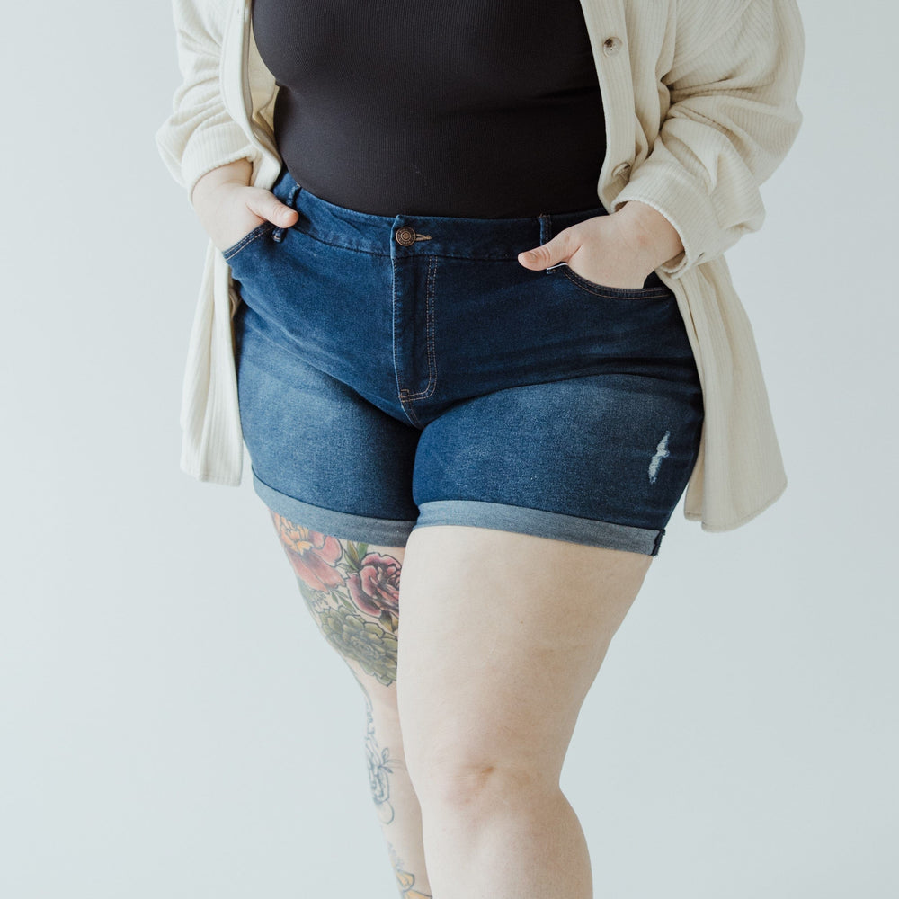 Person in a black top, open beige shirt, and Love Marlow's SLIGHTLY DISTRESSED TUMMY CONTROL CUFFED SHORTS IN DARK WASH with hands in pockets, standing against a plain background. A tattoo is visible on one leg.