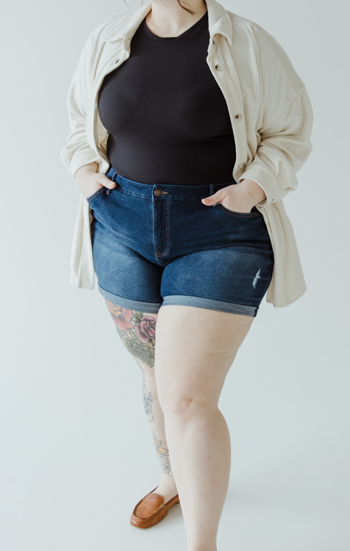 Person in a black top, open beige shirt, and Love Marlow's SLIGHTLY DISTRESSED TUMMY CONTROL CUFFED SHORTS IN DARK WASH with hands in pockets, standing against a plain background. A tattoo is visible on one leg.