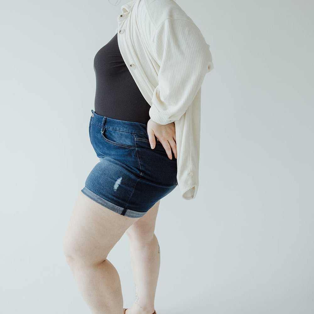 A person stands against a plain background, wearing a cream-colored cardigan over a black top, SLIGHTLY DISTRESSED TUMMY CONTROL CUFFED SHORTS IN DARK WASH by Love Marlow, and brown shoes. They are looking to the side with their hands resting on their hips.