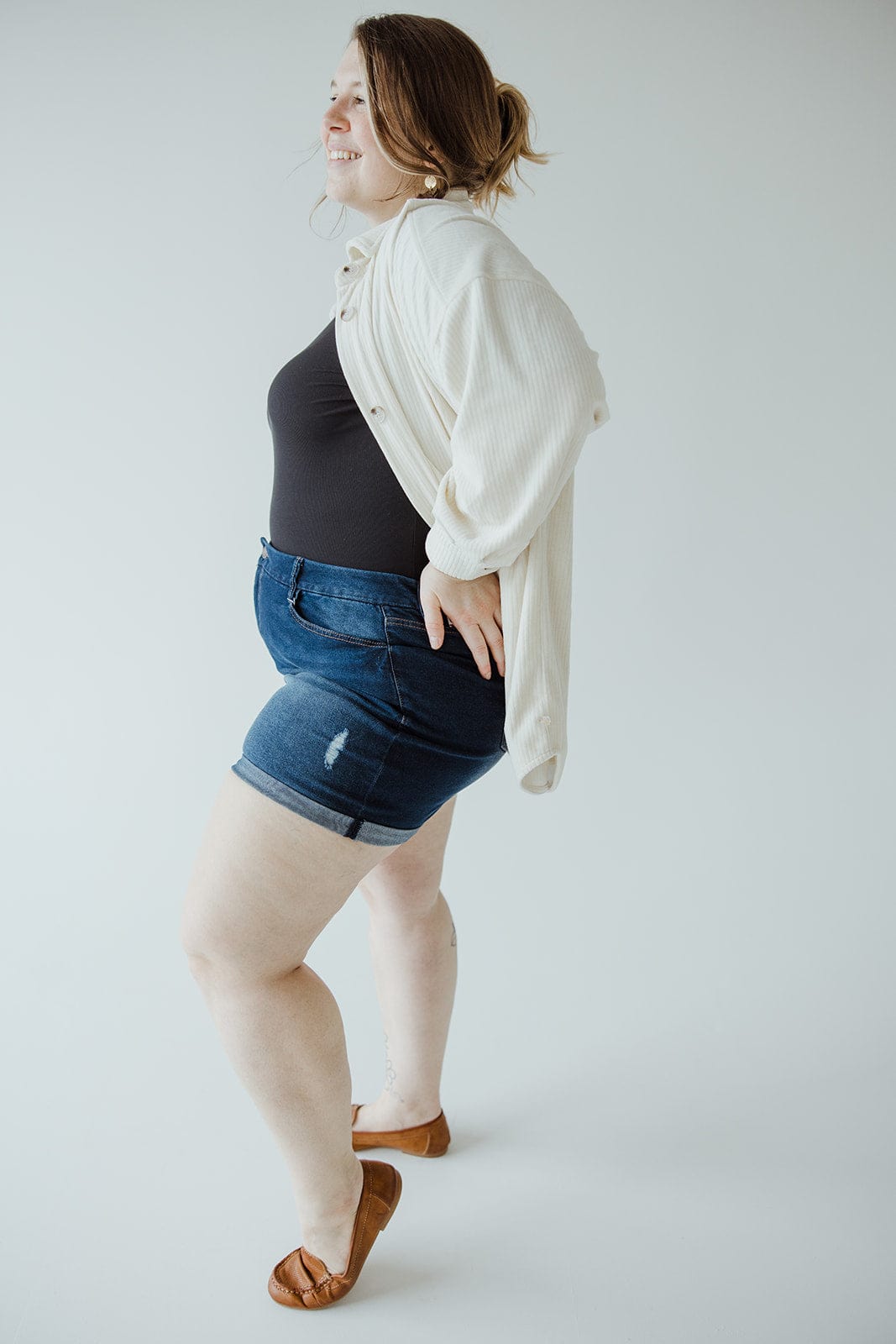 A person stands against a plain background, wearing a cream-colored cardigan over a black top, SLIGHTLY DISTRESSED TUMMY CONTROL CUFFED SHORTS IN DARK WASH by Love Marlow, and brown shoes. They are looking to the side with their hands resting on their hips.