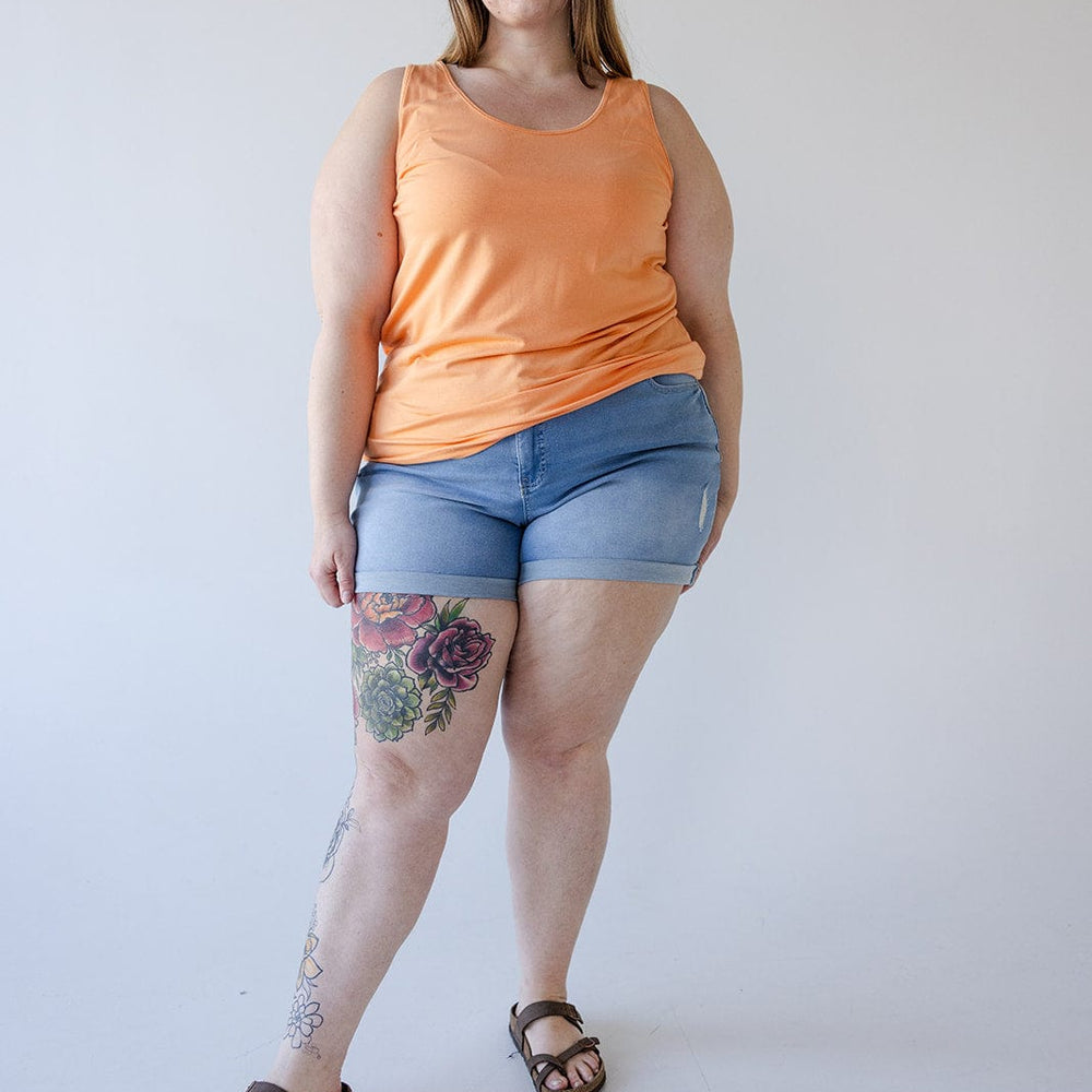 A person stands confidently in a peach tank top, Love Marlow SLIGHTLY DISTRESSED TUMMY CONTROL CUFFED SHORTS IN LIGHTWASH, and sandals. They have long hair, wear glasses, and have colorful floral tattoos on their right leg. The background is plain and light-colored.