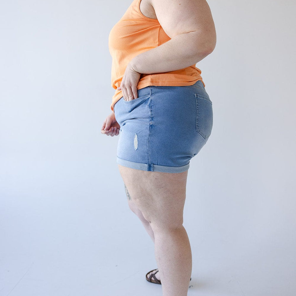 A person wearing an orange tank top, Love Marlow SLIGHTLY DISTRESSED TUMMY CONTROL CUFFED SHORTS IN LIGHTWASH with a small tear, and sandals stands against a plain white background viewed from the side.