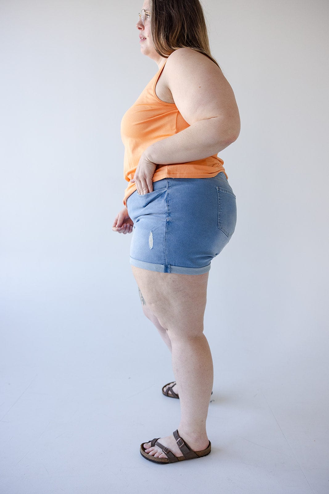 A person wearing an orange tank top, Love Marlow SLIGHTLY DISTRESSED TUMMY CONTROL CUFFED SHORTS IN LIGHTWASH with a small tear, and sandals stands against a plain white background viewed from the side.