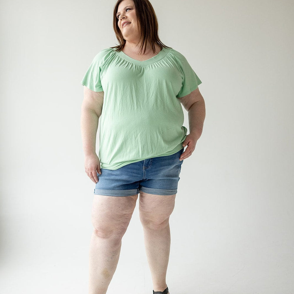 A woman stands against a plain white background, wearing a light green top, SLIGHTLY DISTRESSED TUMMY CONTROL CUFFED SHORTS IN MEDIUM WASH by Love Marlow, and black sandals.