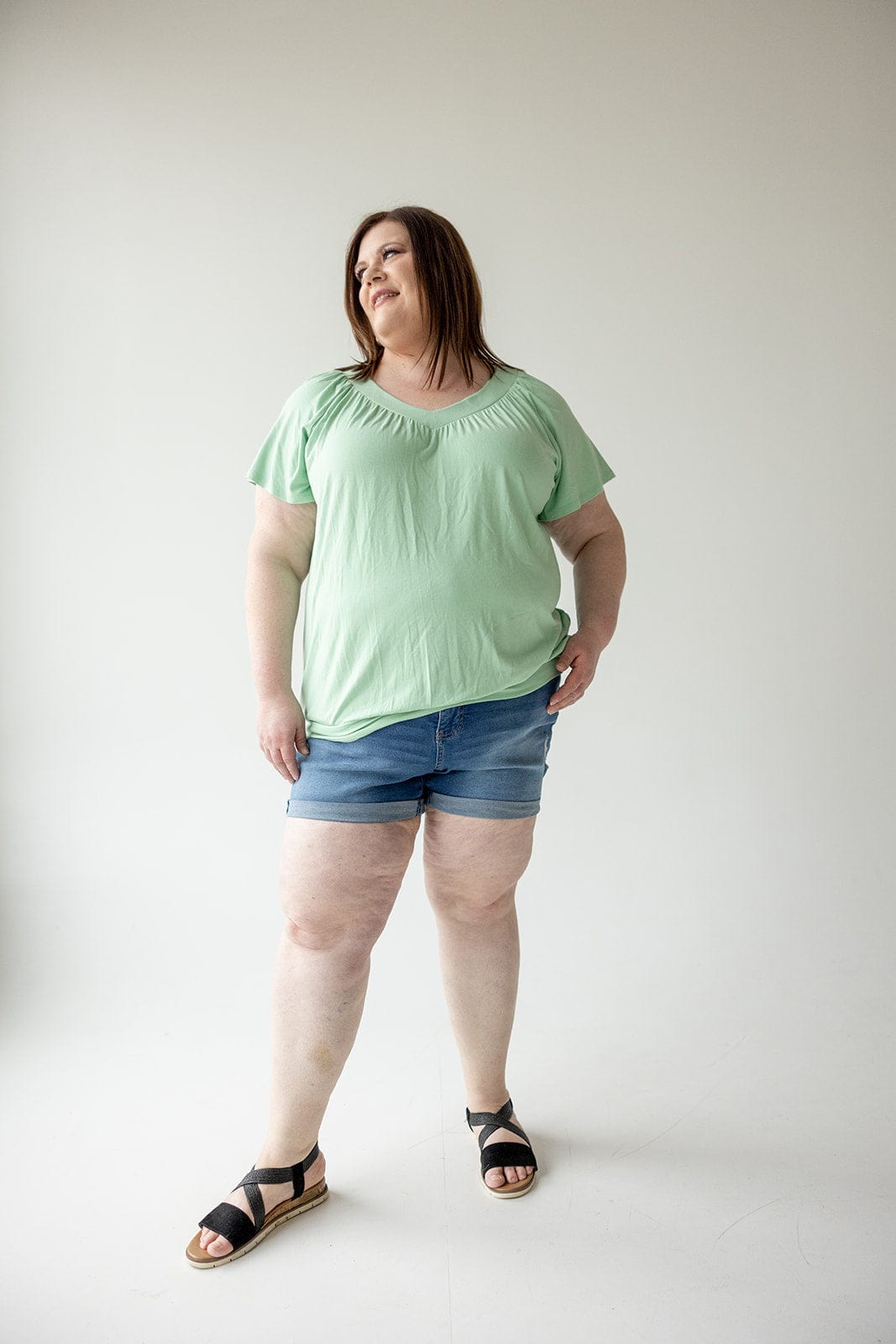 A woman stands against a plain white background, wearing a light green top, SLIGHTLY DISTRESSED TUMMY CONTROL CUFFED SHORTS IN MEDIUM WASH by Love Marlow, and black sandals.