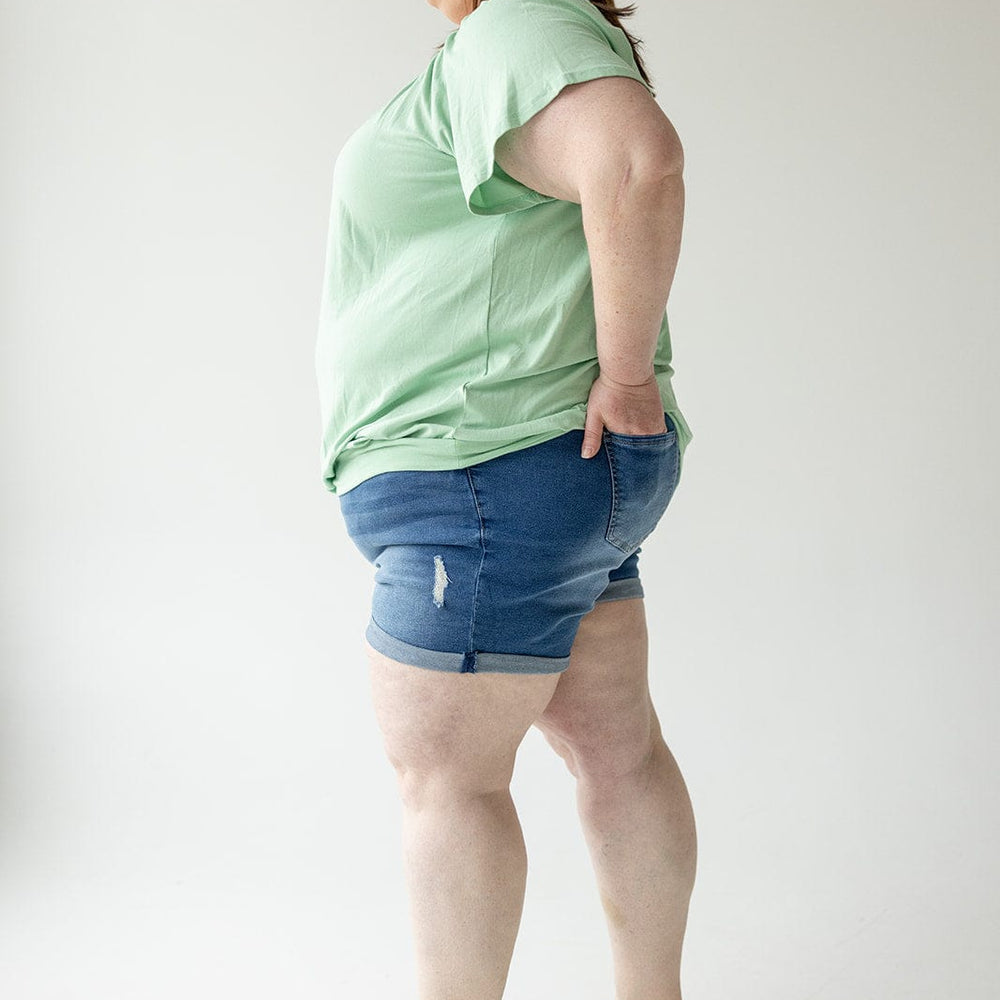 A person stands in profile wearing a green T-shirt, Love Marlow SLIGHTLY DISTRESSED TUMMY CONTROL CUFFED SHORTS IN MEDIUM WASH, and black sandals against a plain white background.