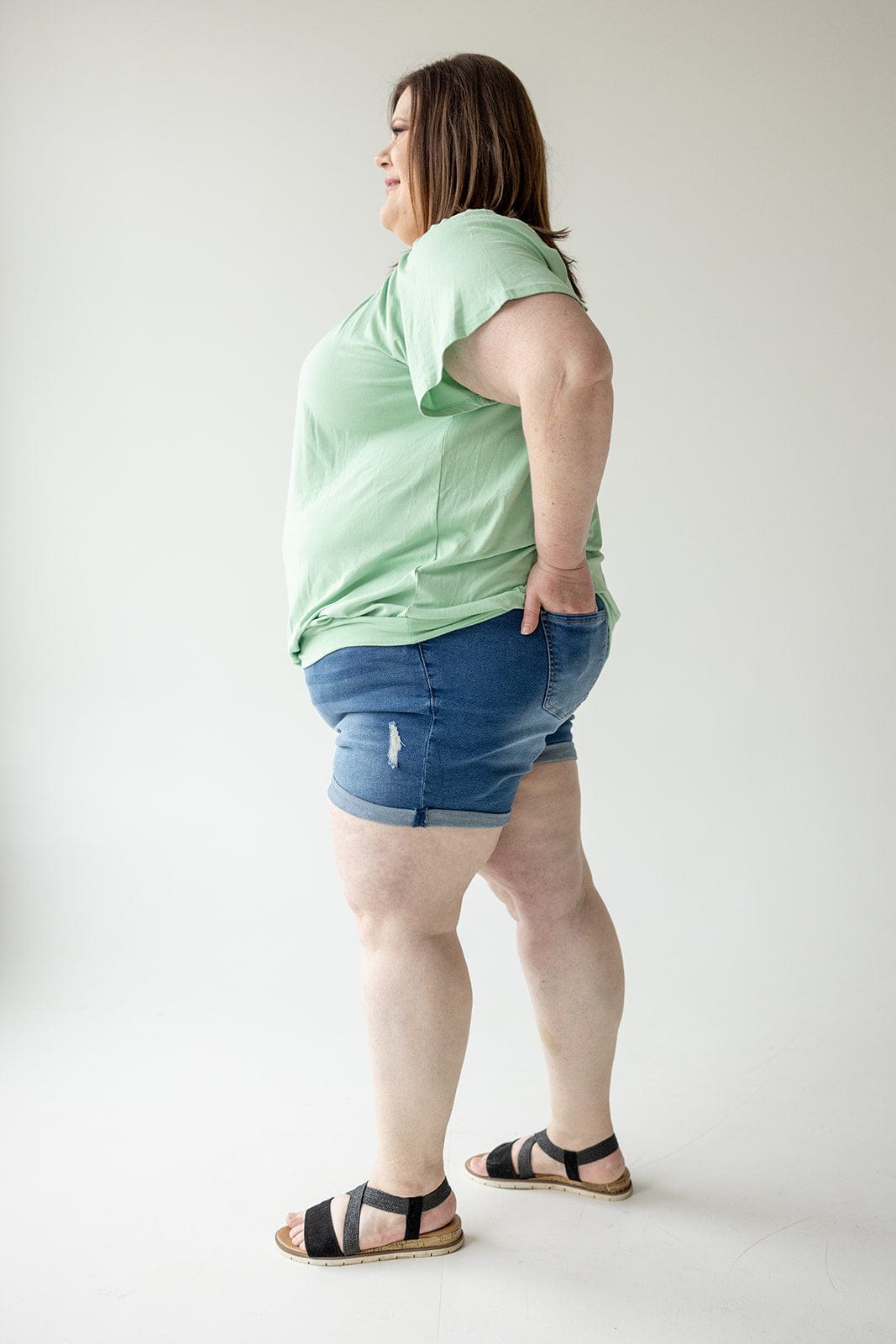 A person stands in profile wearing a green T-shirt, Love Marlow SLIGHTLY DISTRESSED TUMMY CONTROL CUFFED SHORTS IN MEDIUM WASH, and black sandals against a plain white background.