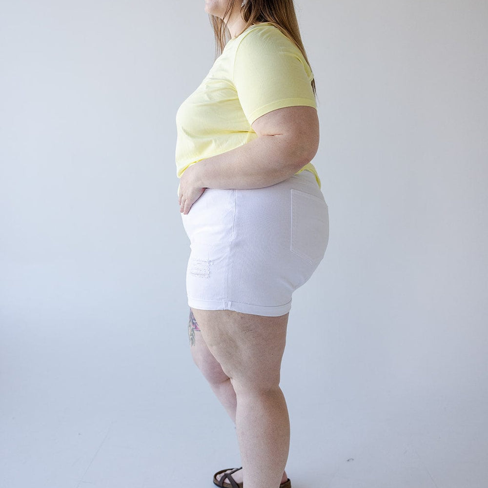 A person stands in a well-lit room, wearing a yellow t-shirt, SLIGHTLY DISTRESSED TUMMY CONTROL CUFFED SHORTS IN WHITE by Love Marlow, and sandals. The person is looking to the left and has a relaxed expression, embodying the perfect summer staple.