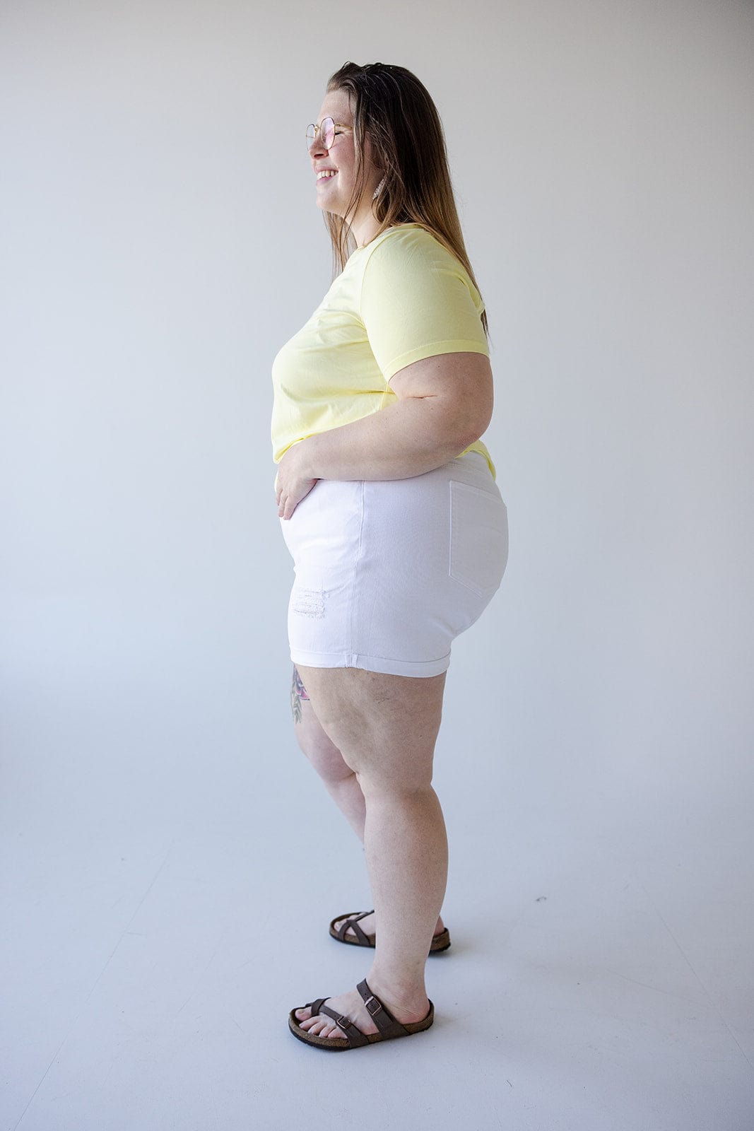 A person stands in a well-lit room, wearing a yellow t-shirt, SLIGHTLY DISTRESSED TUMMY CONTROL CUFFED SHORTS IN WHITE by Love Marlow, and sandals. The person is looking to the left and has a relaxed expression, embodying the perfect summer staple.