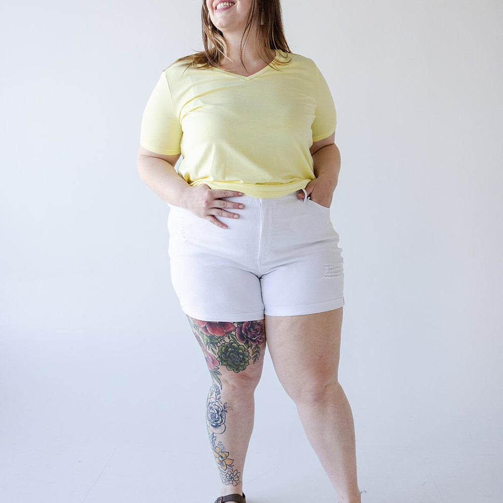 A woman stands against a plain white background wearing a yellow shirt, Love Marlow SLIGHTLY DISTRESSED TUMMY CONTROL CUFFED SHORTS IN WHITE, and brown sandals. She has tattoos on her right leg and smiles while looking slightly to the right.