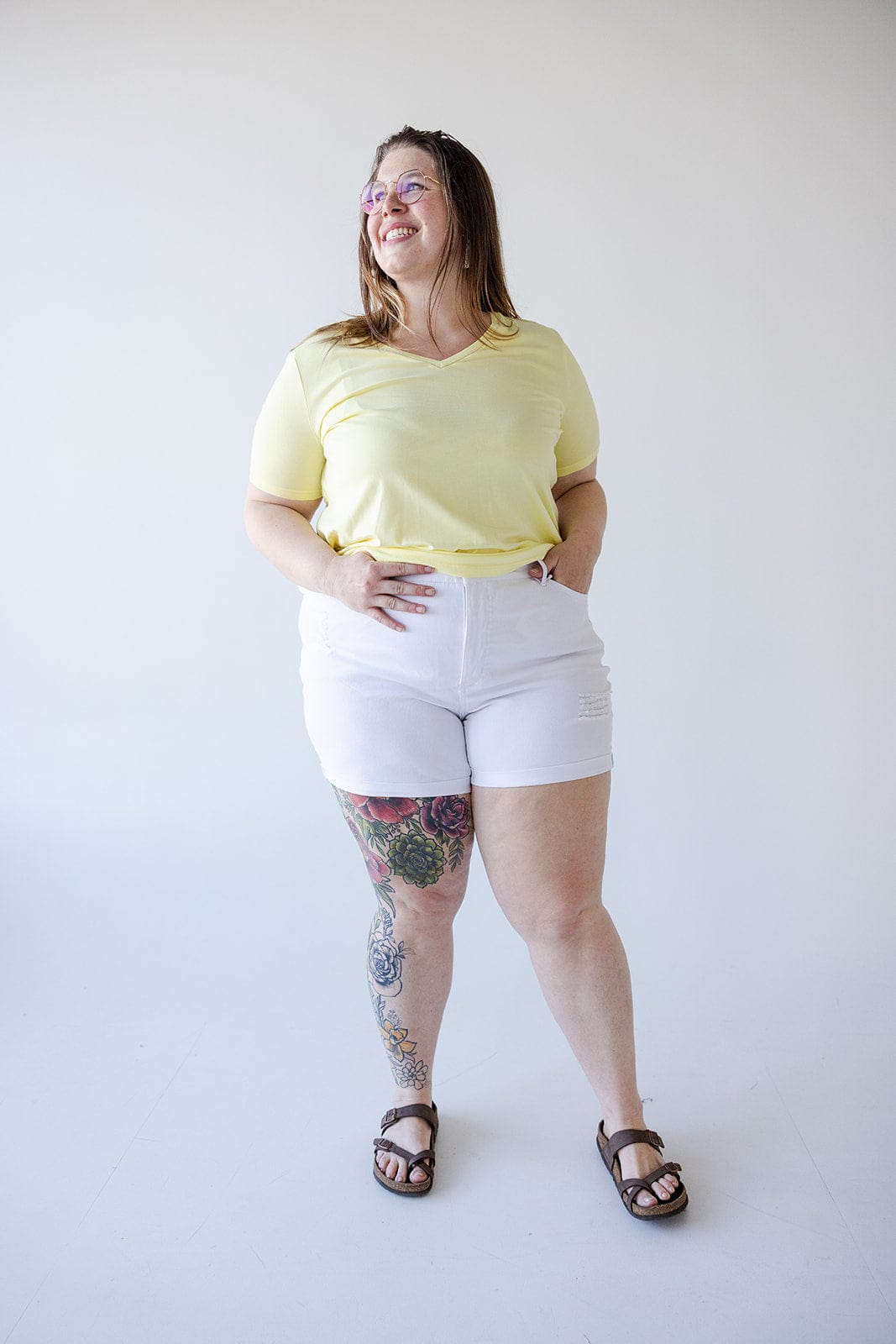 A woman stands against a plain white background wearing a yellow shirt, Love Marlow SLIGHTLY DISTRESSED TUMMY CONTROL CUFFED SHORTS IN WHITE, and brown sandals. She has tattoos on her right leg and smiles while looking slightly to the right.