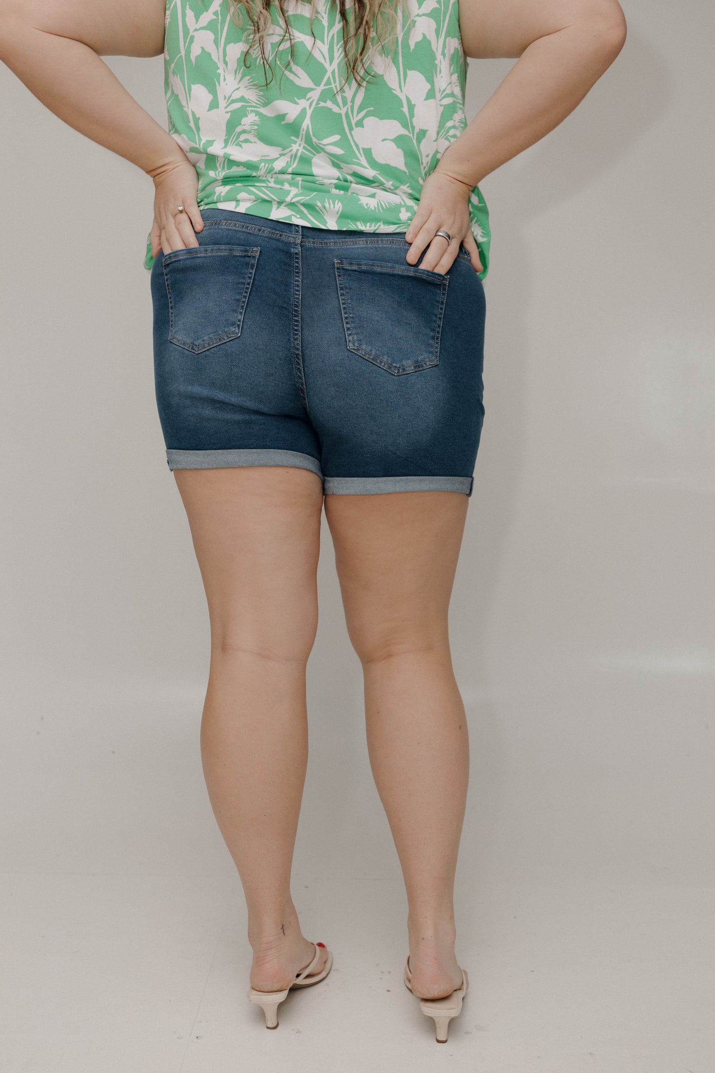 Person wearing a green floral top and blue denim shorts on a plain background