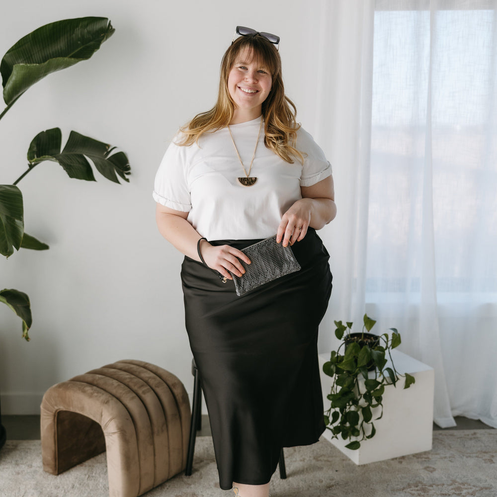 Woman in a white top and black skirt standing in a room with plants and a brown ottoman. Beaded Wallet clutch.