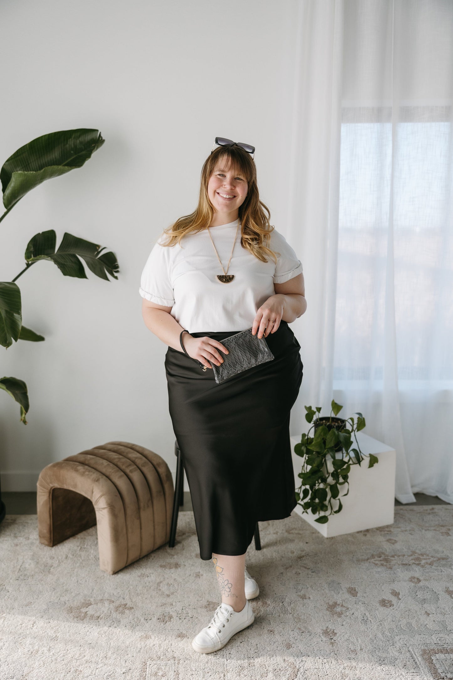 Woman in a white top and black skirt standing in a room with plants and a brown ottoman. Beaded Wallet clutch.