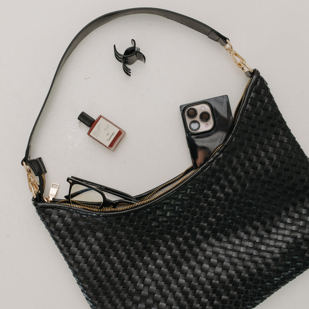 Black woven handbag with items like a phone, sunglasses, and nail polish on a light gray background