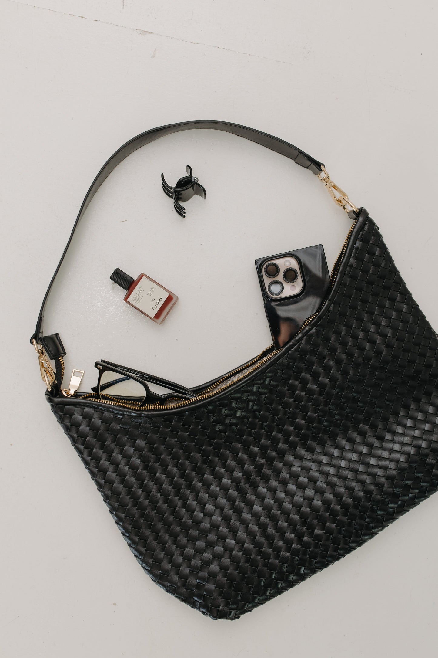 Black woven handbag with items like a phone, sunglasses, and nail polish on a light gray background